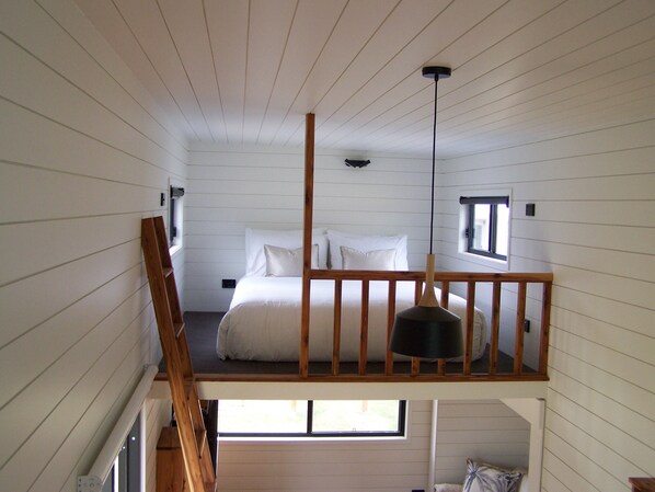 A bedroom with a loft bed, a wooden railing, a hanging light fixture, and a window providing natural light.