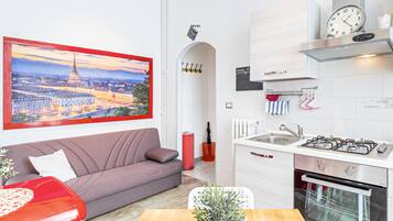 A modern kitchen with a gas stove, a sink, and a stainless steel oven. There is a red couch and a table with a potted plant and cups.