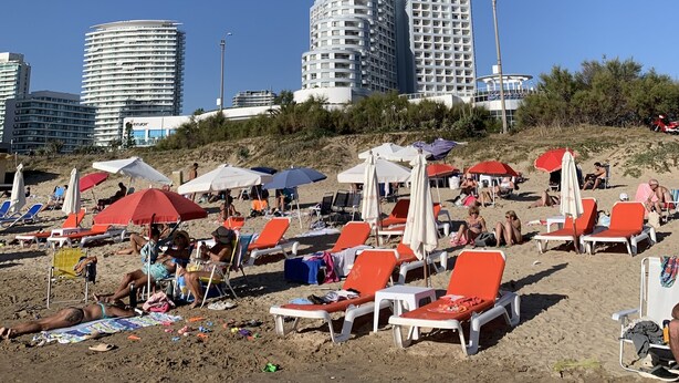 A beach with rows of orange lounge chairs and umbrellas, a clear blue sky, and high-rise buildings in the background. - Lindo apartamento a una cuadra de la Playa Brava. (Punta del Este)