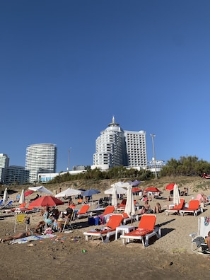 Een strand met rijen oranje ligbedden en parasols, een strakblauwe lucht en hoge gebouwen op de achtergrond.