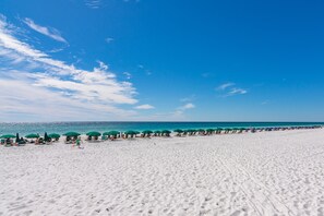 Une plage bordée de rangées de parasols verts et de chaises longues, un ciel bleu azur et l’océan en toile de fond.