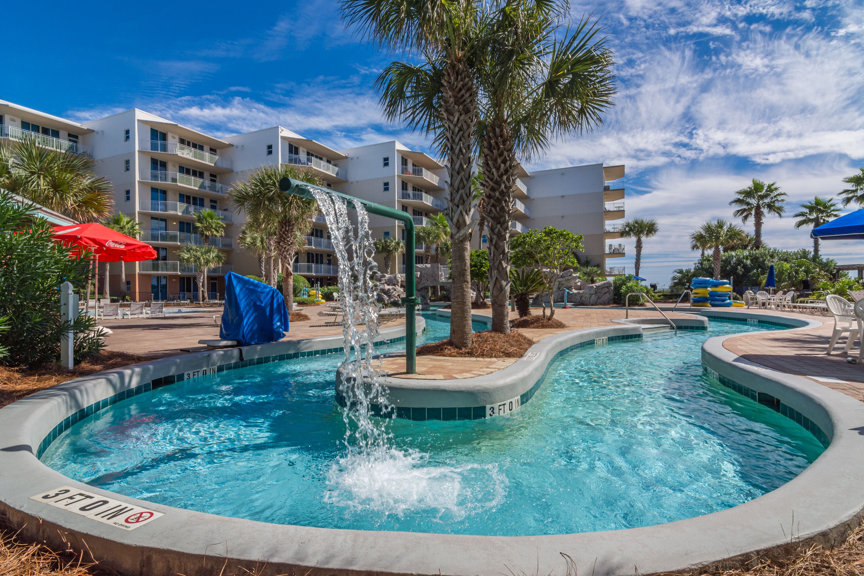 A swimming pool with a waterfall feature, surrounded by palm trees and a multi-story building with balconies.