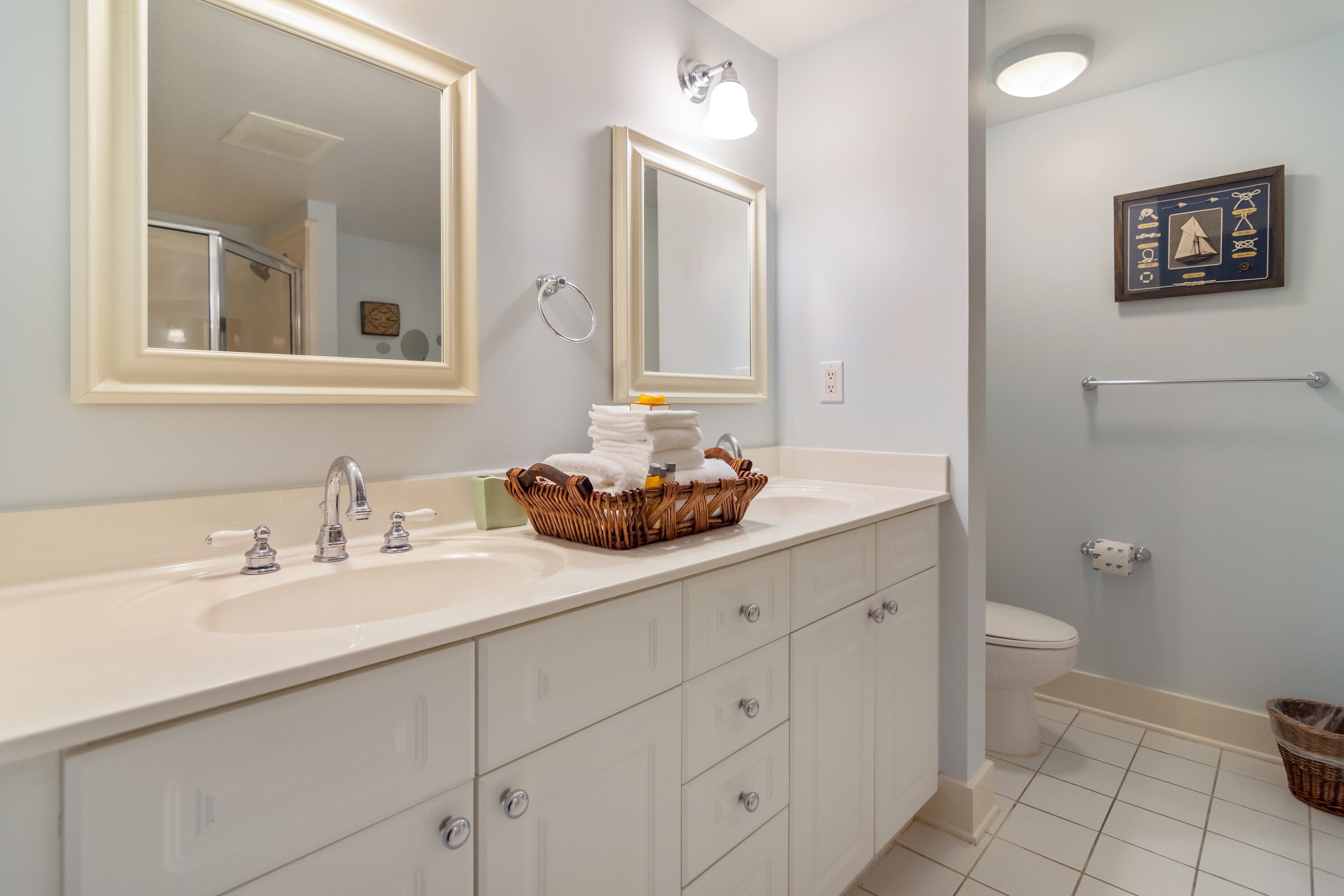 Une salle de bain avec un lavabo blanc, deux miroirs, un porte-serviettes et un tableau encadré au mur.