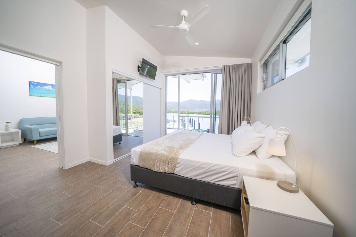A modern bedroom with a large bed, a ceiling fan, a TV, and a balcony with a view of the mountains.