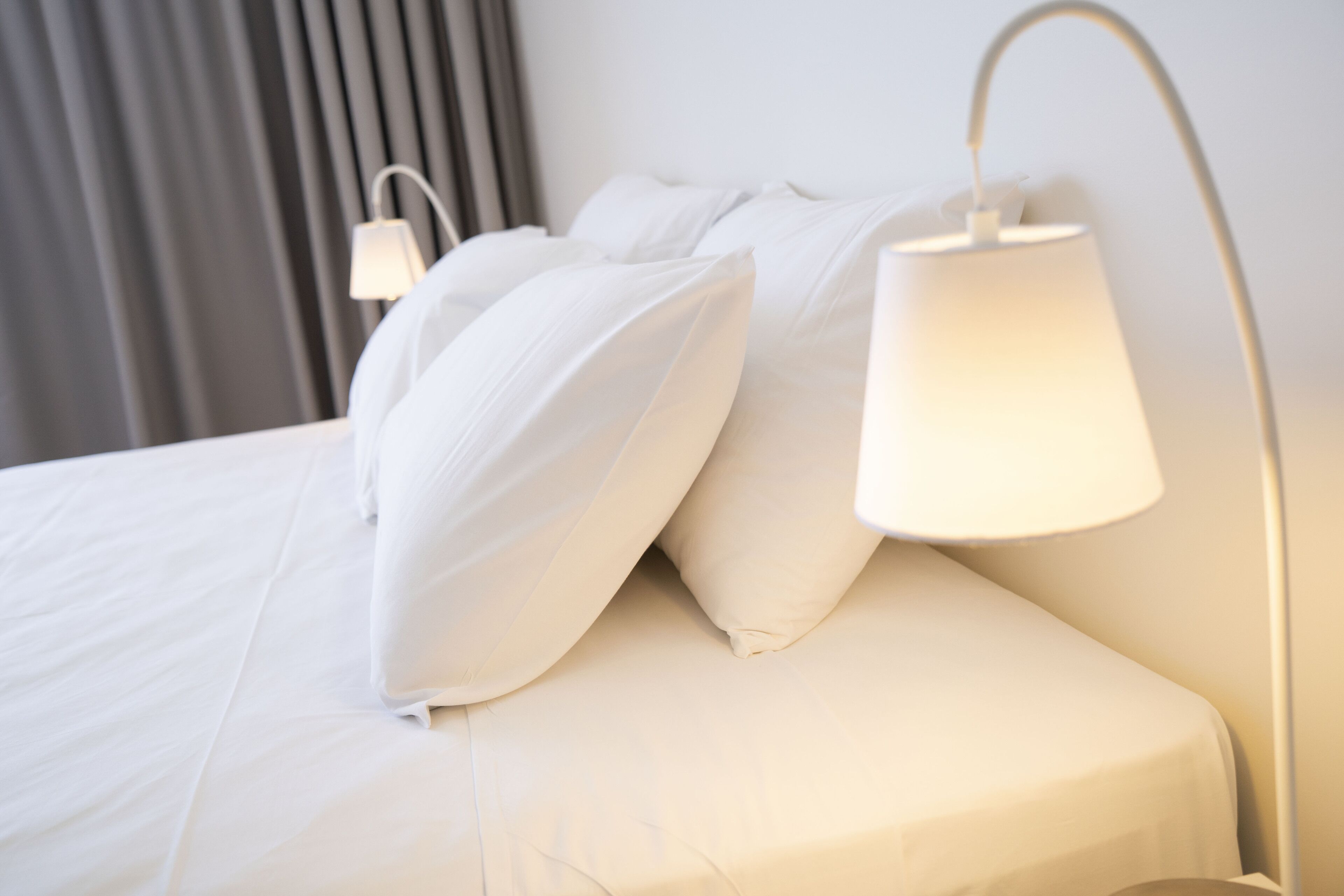 Una cama bien hecha con sábanas y almohadas blancas, una lámpara de noche y un aplique de pared.
