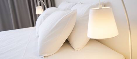 Cama bem arrumada com lençóis e almofadas brancos, luminária de mesa e luminária na parede.