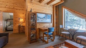 Interior acolhedor de uma cabana com paredes de madeira, área de jantar com mesa e cadeiras, estante de livros e janela com vista nevada.