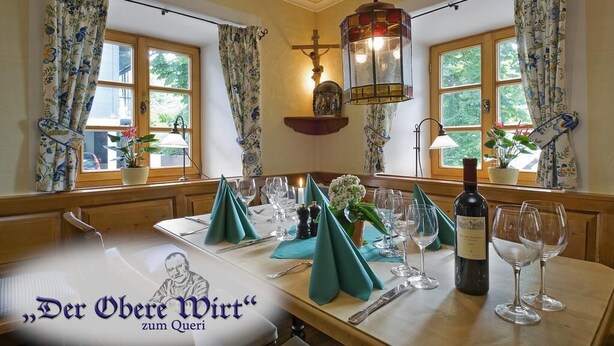 A dining table set with green napkins, wine glasses, and a bottle of wine, with floral curtains and a crucifix on the wall. - Der Obere Wirt zum Queri (Andechs)
