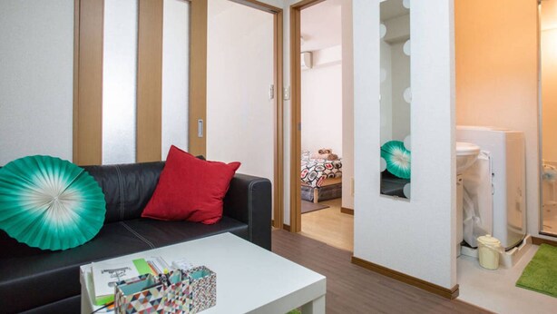 A modern living room with a black leather sofa, a red pillow, and a white coffee table. There is a door leading to a bedroom with a bed and a bathroom visible. - Not Ninja Osaka Namba 1 (Osaka)
