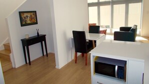 A modern living room with a white sofa, a black chair, and a dining table. There is a staircase on the left and a window with blinds on the right.
