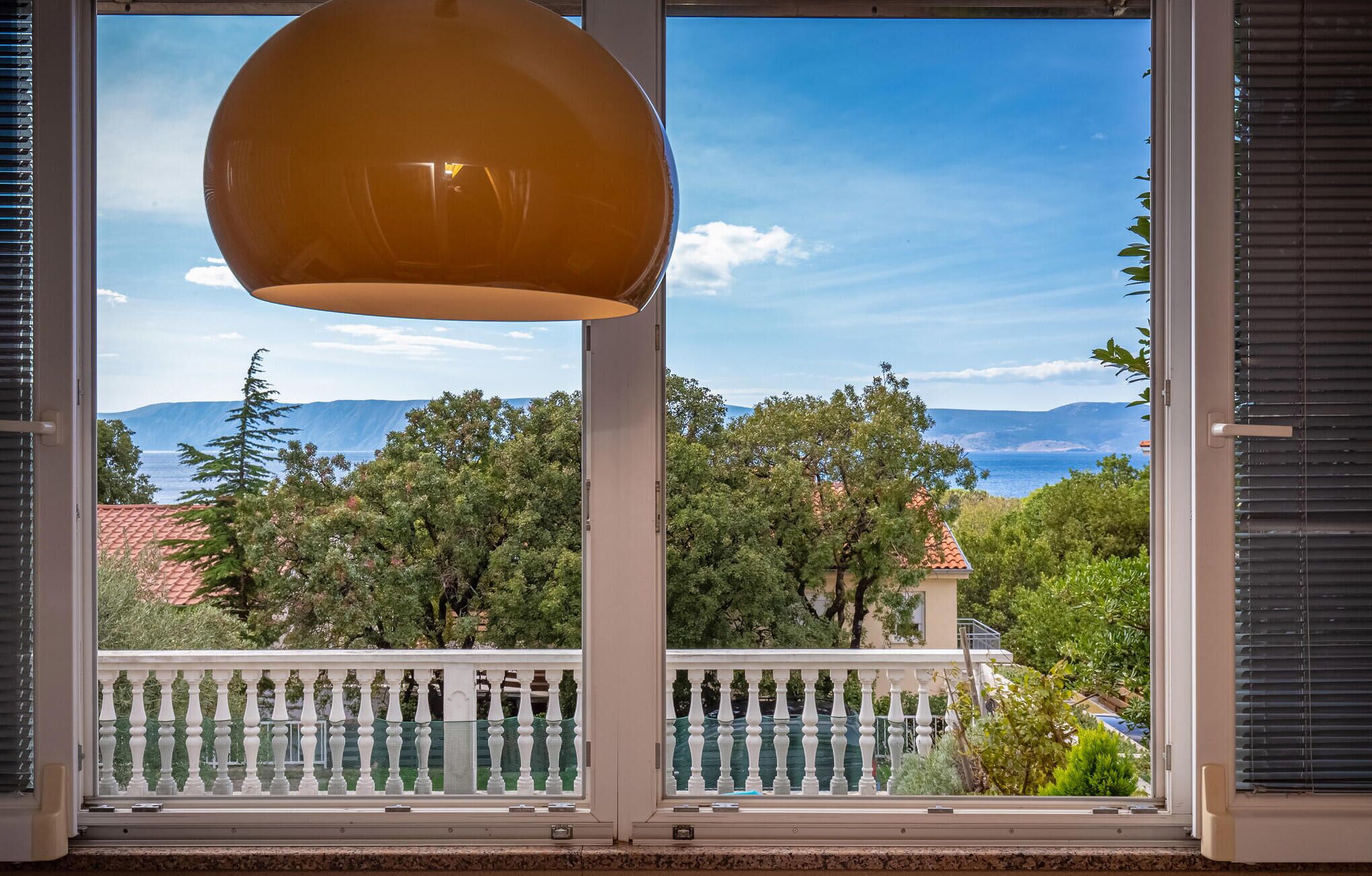Een balkon met een witte balustrade, een hanglamp en een uitzicht op een bergachtig landschap door een raam met zonwering.