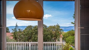 Ein Balkon mit weißer Brüstung, einer hängenden Pendelleuchte und Blick auf eine Berglandschaft durch ein Fenster mit Jalousien.