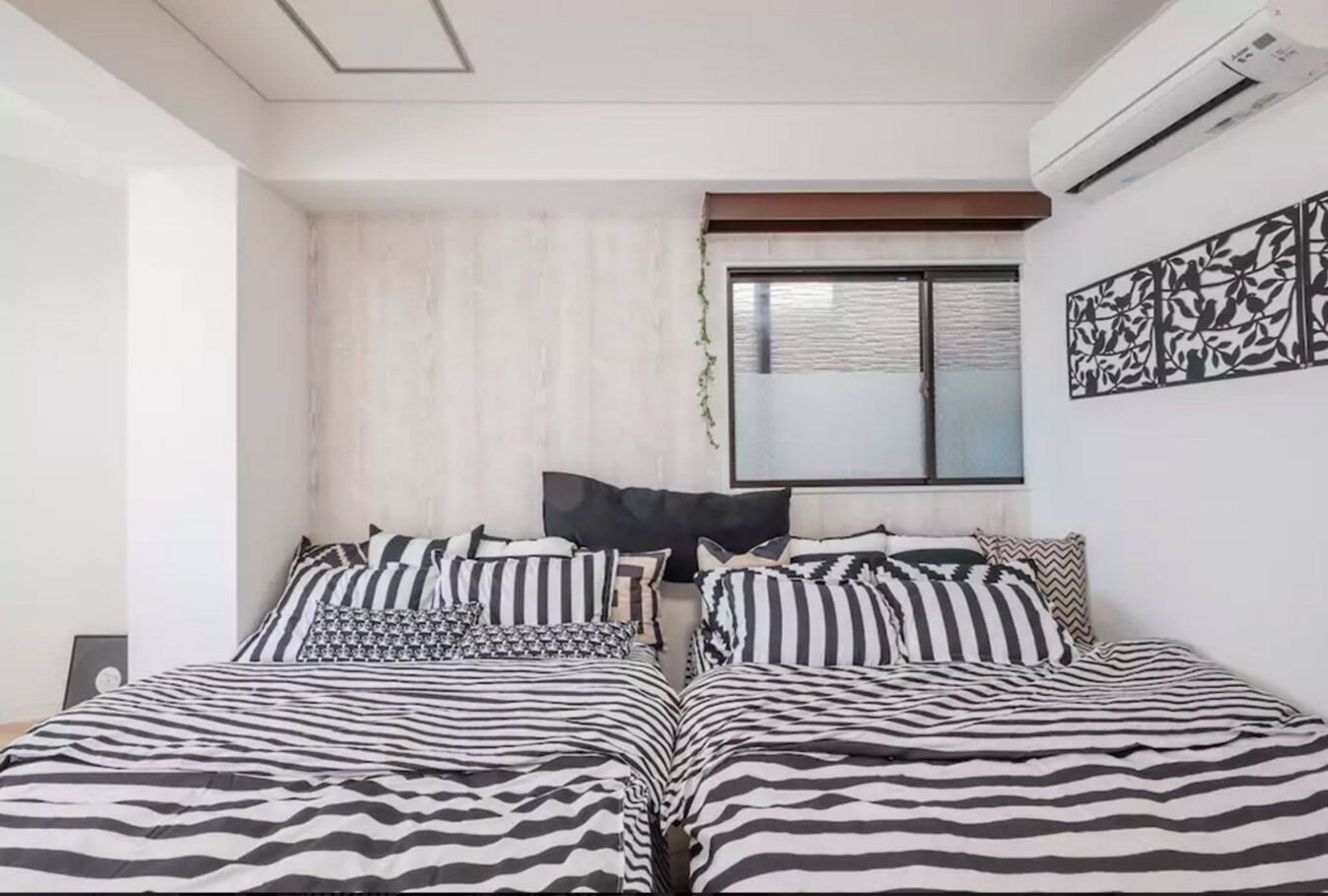 Une chambre avec deux lits, une literie à rayures, une fenêtre avec des stores, un climatiseur et une œuvre d’art accrochée au mur.