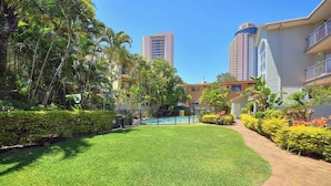 A well-maintained courtyard with a swimming pool, surrounded by lush greenery and tall buildings in the background. - Beautiful 2 Bedroom Retreat  ( 1 min from tramline) (Surfers Paradise)