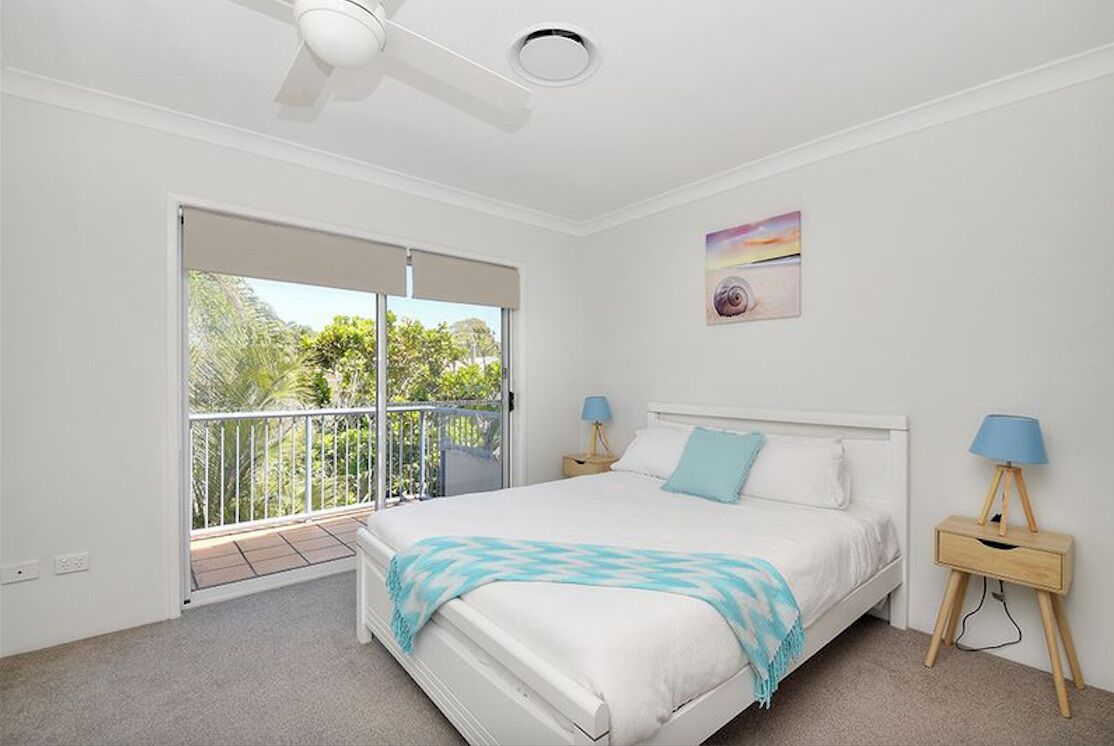 A bedroom with a bed, a ceiling fan, a nightstand with a lamp, and a balcony with a view of greenery.