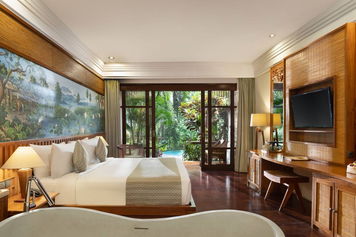 A spacious hotel room with a large bed, a desk, a television, and a view of a lush garden.