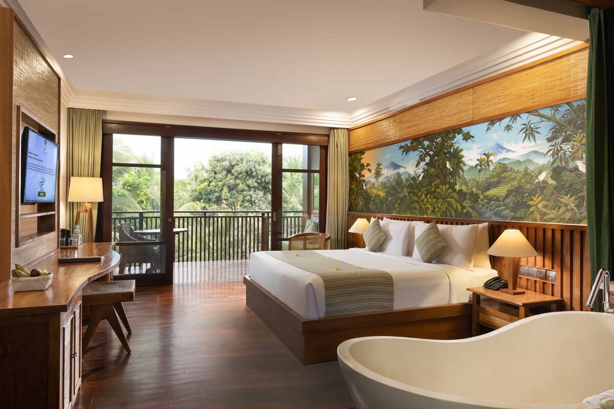 A hotel room with a large bed, a bathtub, a desk with a computer, and a balcony with a view of the outdoors.