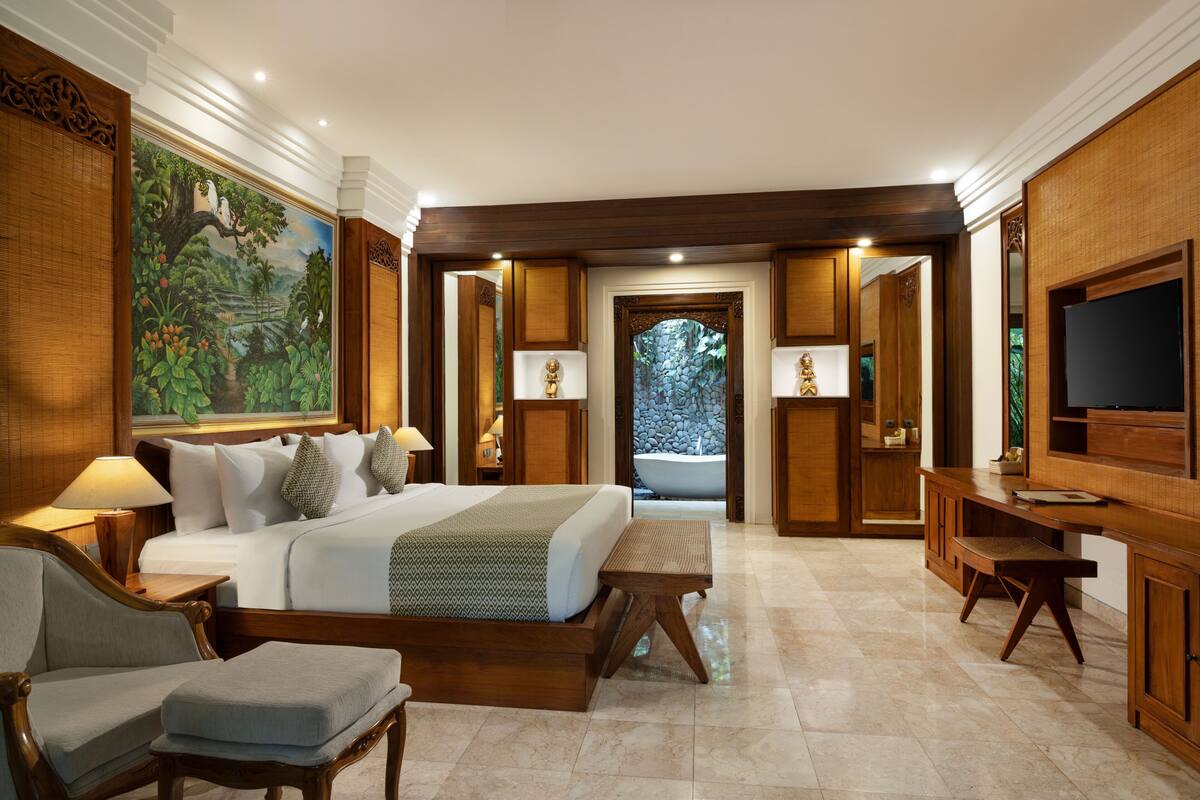 A spacious bedroom with a large bed, a sitting area, a desk, and a television. There is a large painting on the wall and a view of a garden through the door.