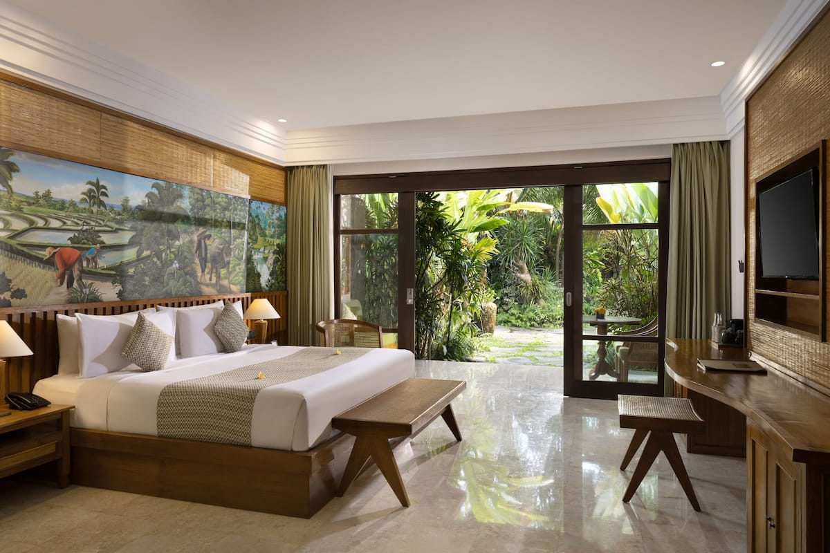 A spacious bedroom with a large bed, a wooden nightstand, a bedside table, a TV, and a view of a lush garden through a glass door.