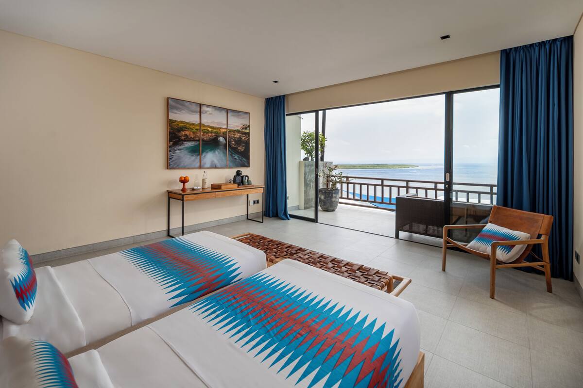 A hotel room with a large bed, a desk, a chair, and a balcony with a view of the sea.