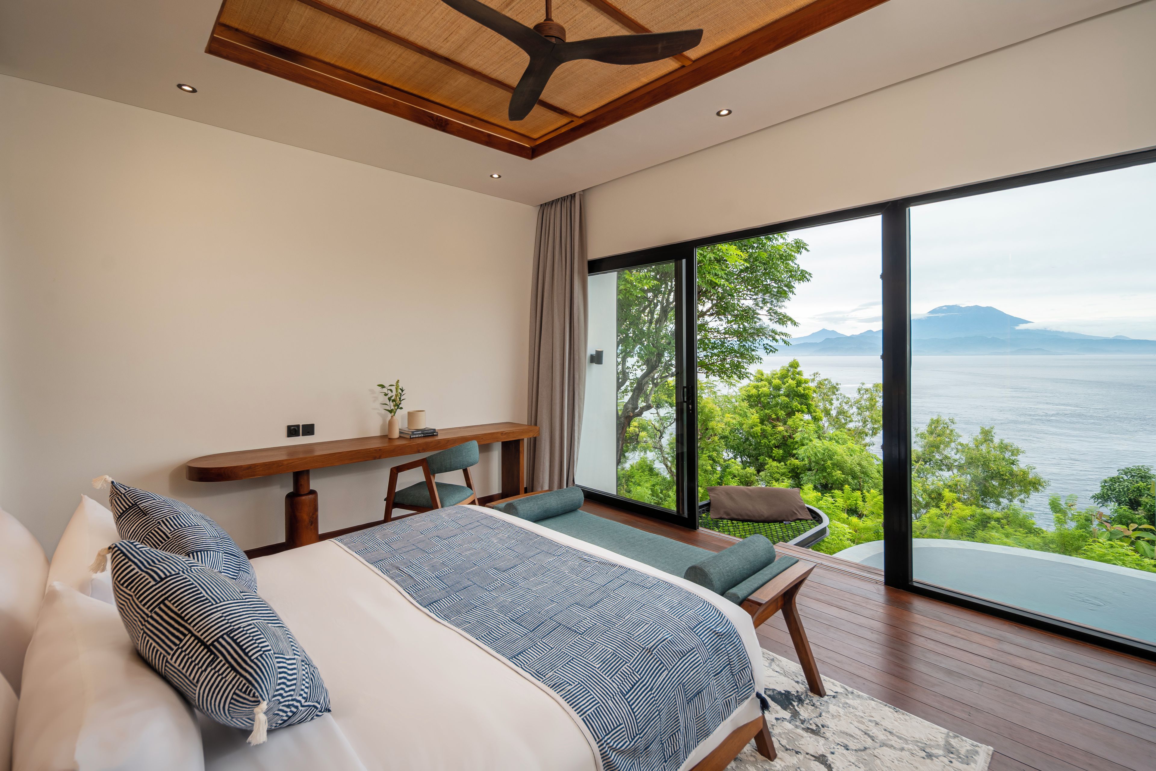 A bedroom with a large window, a bed, a desk, and a view of the sea.