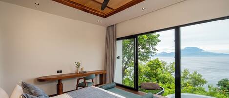 A bedroom with a large window, a bed, a desk, and a view of the sea.