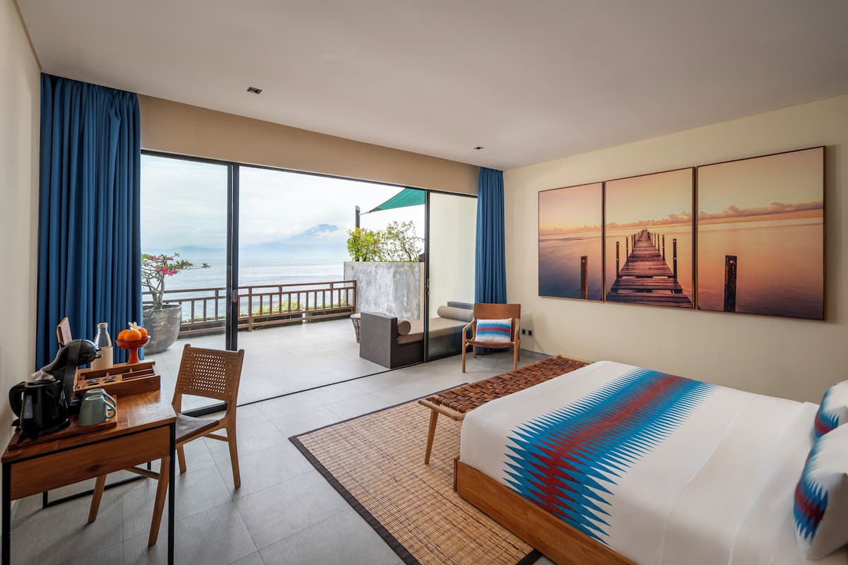 A hotel room with a bed, a desk, a chair, a view of the sea, and a sunset painting.