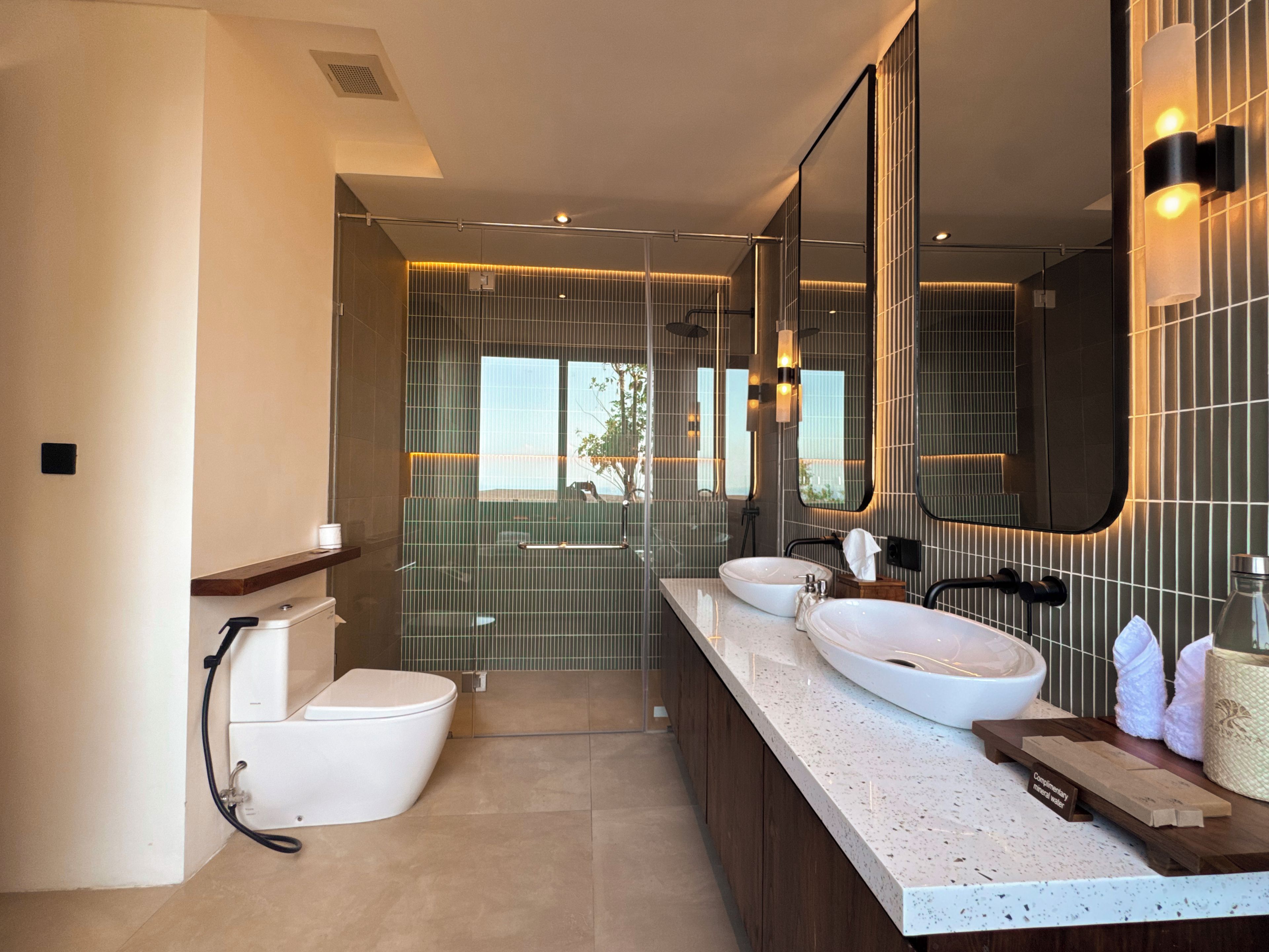 A modern bathroom with a large mirror, two sinks, a toilet, and a shower area.