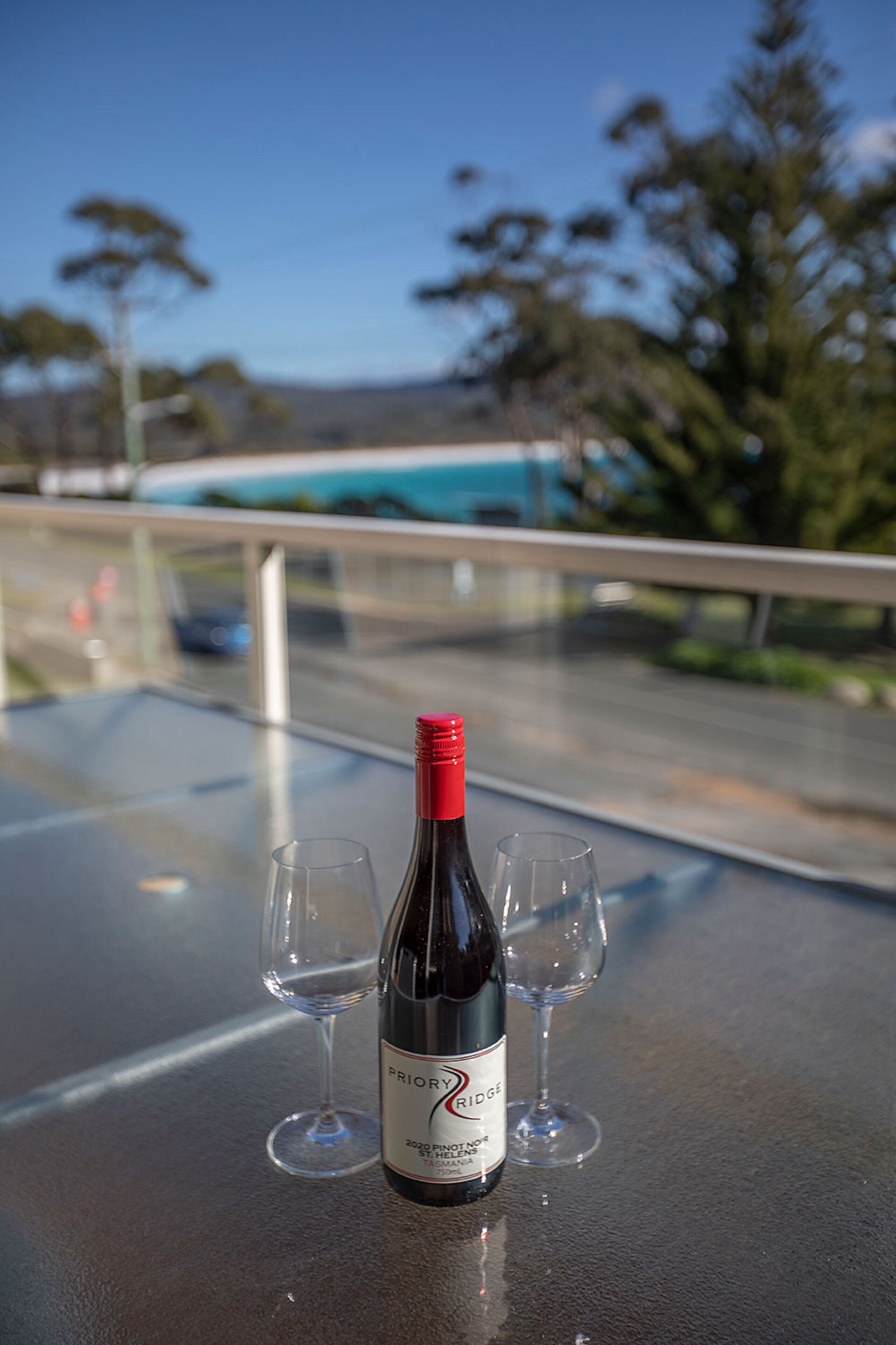 A bottle of wine and two glasses on a table with a view of trees and a road in the background.