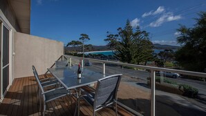 Un balcon avec une table et des chaises, donnant sur une route bordée d’arbres et où circulent des voitures.