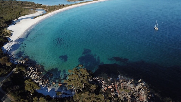Vue aérienne d’une zone côtière avec une plage de sable, des eaux d’un bleu limpide et un voilier.