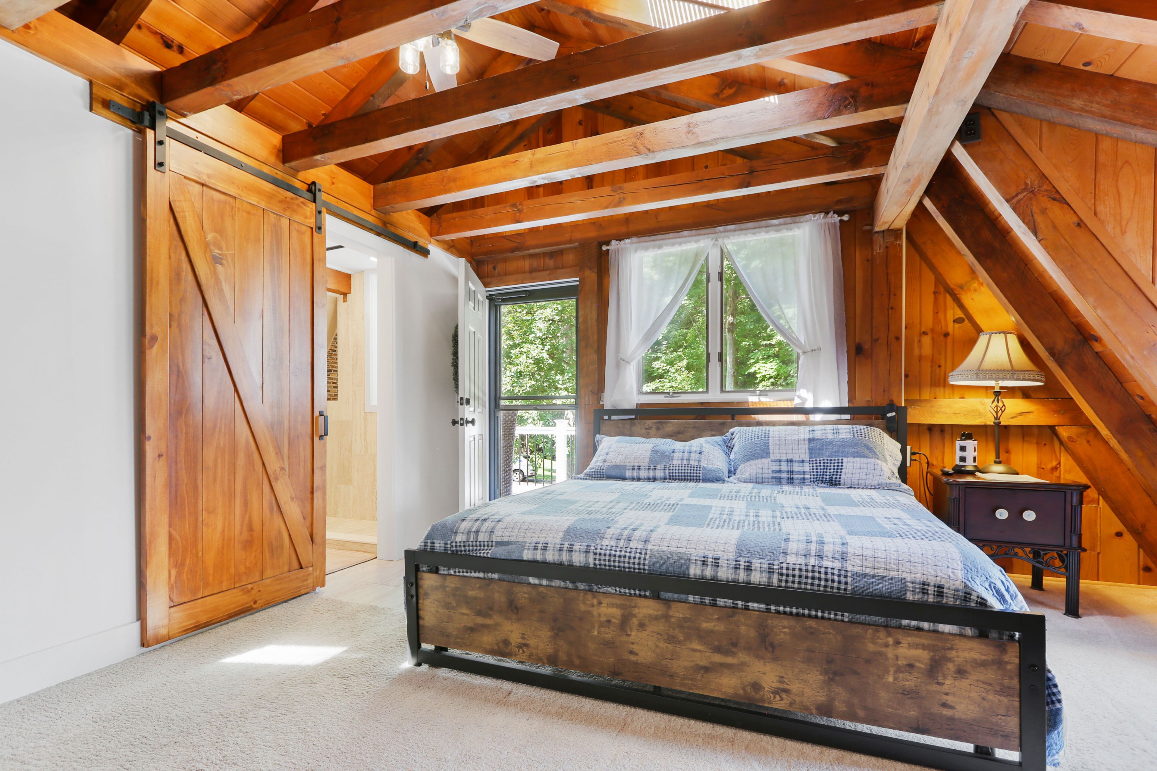Ein Schlafzimmer mit Holzboden, einem Bett mit kariertem Überwurf, einem Nachttisch mit Lampe und einem Fenster mit Vorhängen.