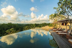Una piscina infinita con sillones de madera, una pared de piedra y exuberante vegetación al fondo.