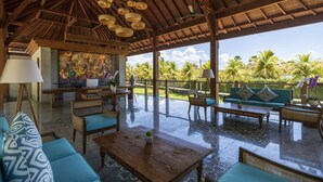 A spacious outdoor lounge area with wooden furniture, a thatched roof, and a view of palm trees. - Adiwana Bisma (Ubud)