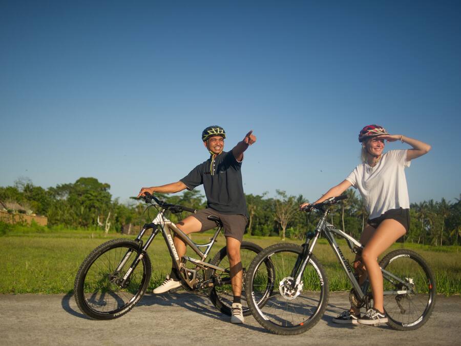 Two people on bicycles on a paved path with greenery in the background.