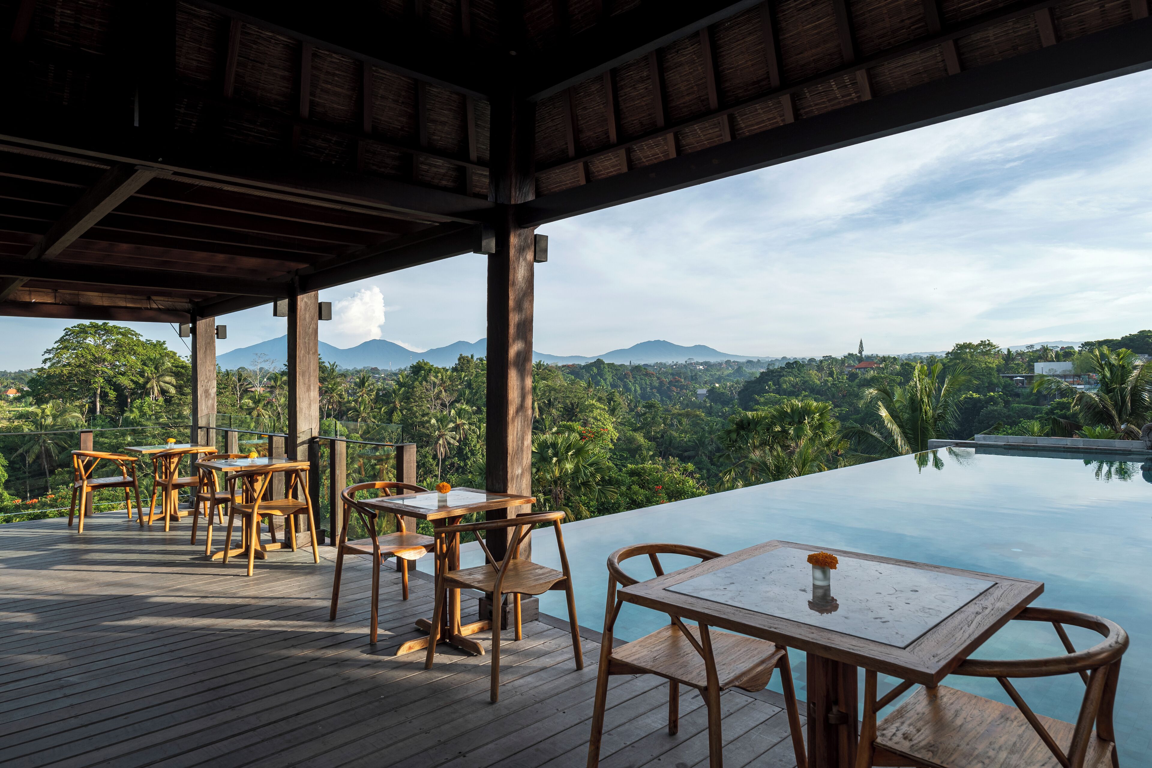 An outdoor dining area with wooden tables and chairs, overlooking a pool and a scenic view of mountains and greenery.