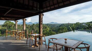 An outdoor dining area with wooden tables and chairs, overlooking a pool and a scenic view of mountains and greenery.