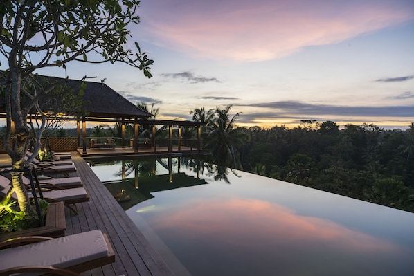 Una piscina infinita tranquila con tumbonas de madera en la terraza, un área de estar cubierta y abundante vegetación tropical.