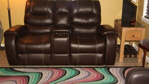 Un canapé en cuir marron assorti d’un pouf, un tapis rayé aux couleurs vives, une table d’appoint en bois avec un téléviseur, et un miroir au-dessus du canapé.