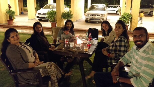 A group of people seated at an outdoor table with a view of parked cars and a building in the background. - Alaya Stays Veera Strand Park (Calangute)