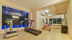 A gym with treadmills, an elliptical, and a stationary bike, with a pool visible through the window. - Alaya Stays Veera Strand Park (Calangute)