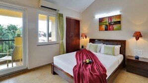 A hotel room with a bed, a nightstand, a lamp, a window with curtains, a balcony with a chair, and a painting on the wall. - Alaya Stays Veera Strand Park (Calangute)
