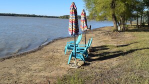 Escena junto a un lago con sillas azules y sombrillas a rayas cerca de la orilla.