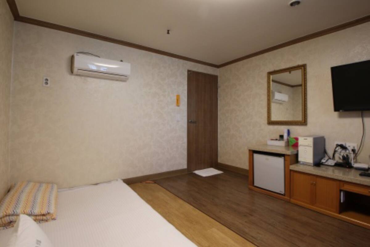 A hotel room with a bed, a small desk with a mirror, a television, and an air conditioning unit.