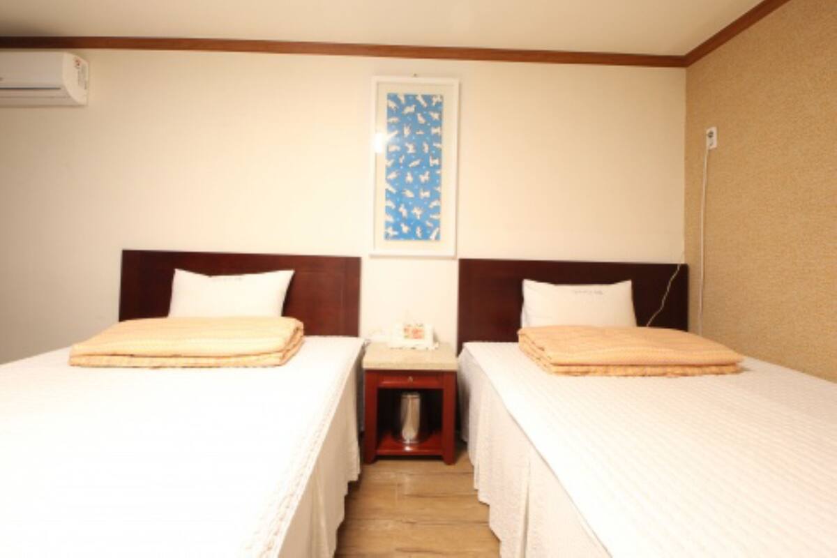A hotel room with two single beds, a small bedside table with a lamp, a framed picture on the wall, and a wooden floor.