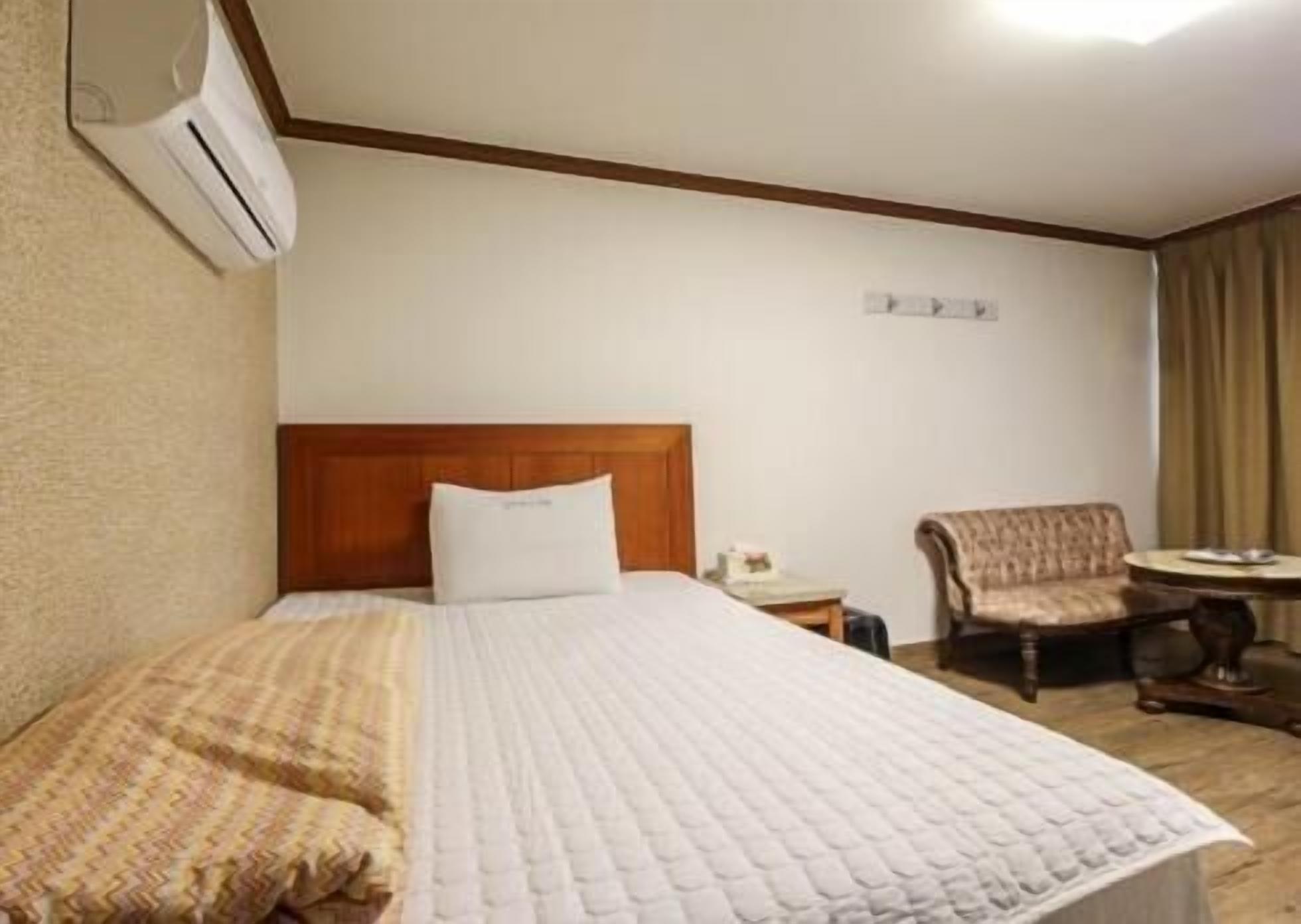 A hotel room with a bed, a chair, a small table, and an air conditioning unit.