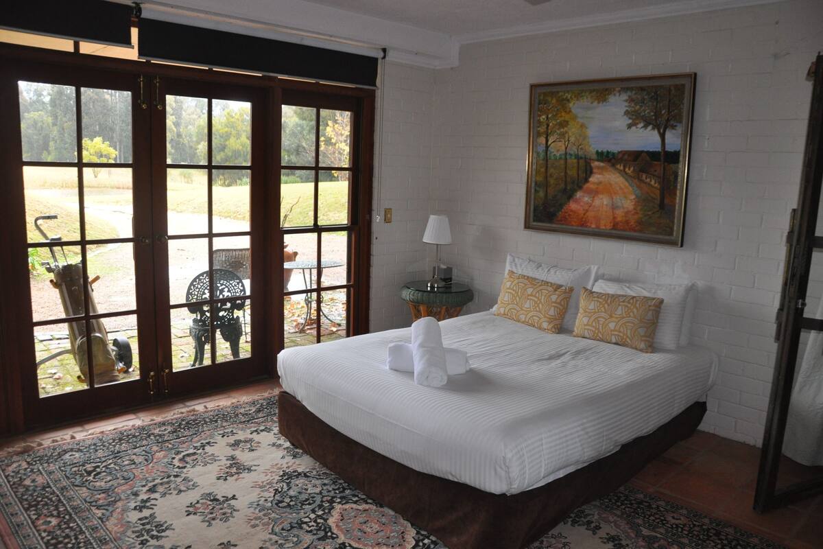 A bedroom with a large bed, a painting on the wall, and a view through a glass door.