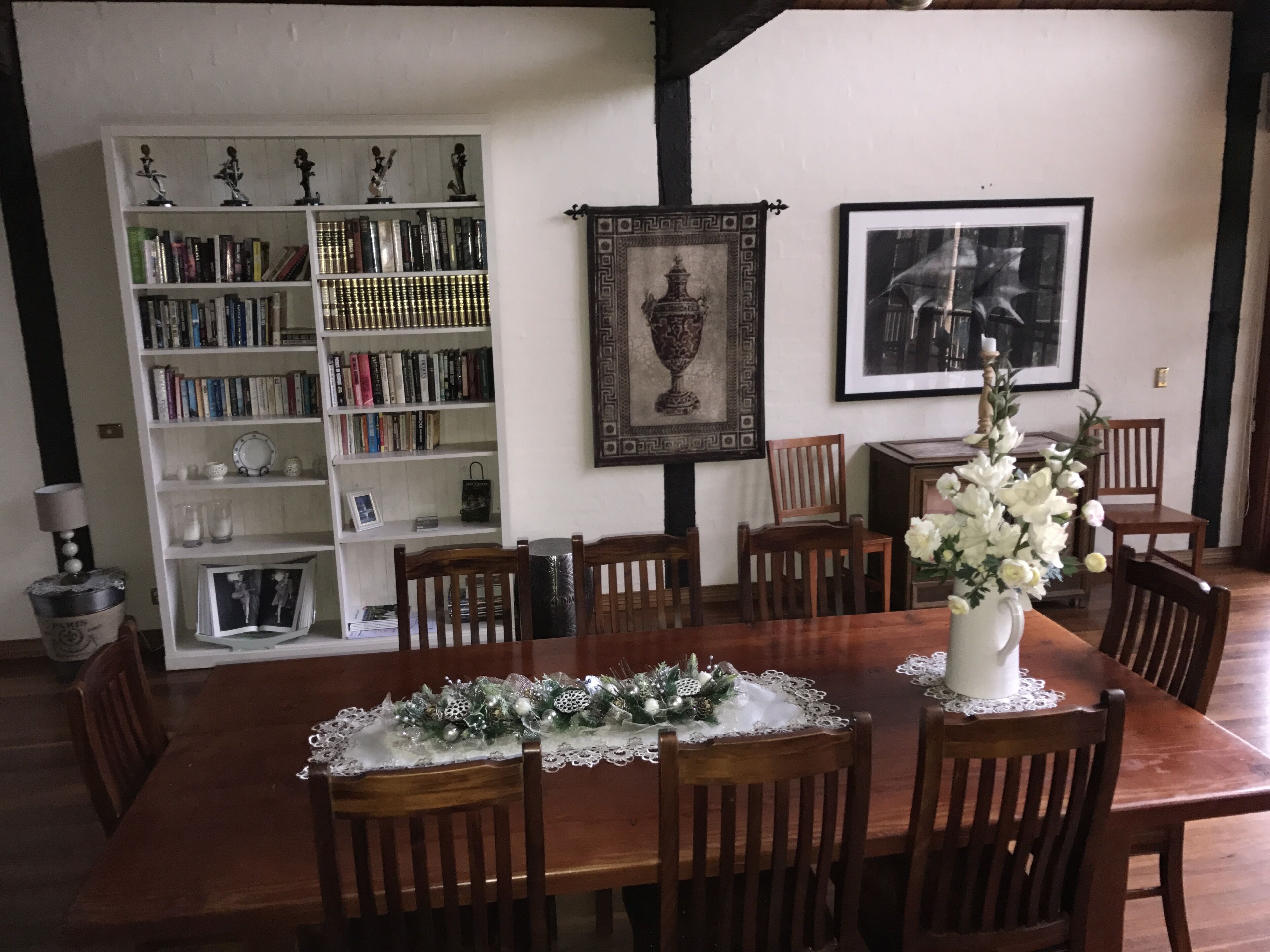 A dining area with a wooden table set for six, surrounded by wooden chairs. A white vase with flowers is on the table.