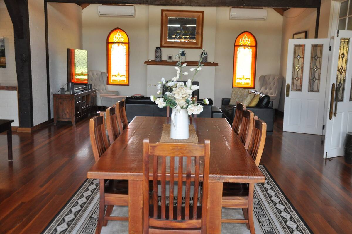 A dining room with a wooden table and chairs, a vase with flowers, and two stained glass windows.