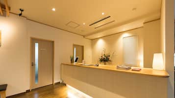 A reception area with a wooden reception desk, a potted plant, a flower arrangement, and a window with blinds.